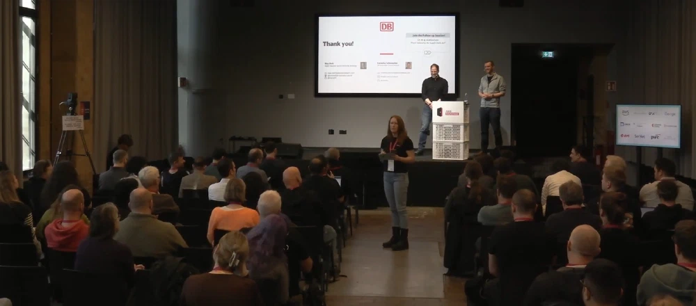 Cornelius Schumacher and Max Mehl giving the presentation at FOSS Backstage 2026. It's a total view of the auditorium from the back, with the two speakers on stage and the final slide in the background.